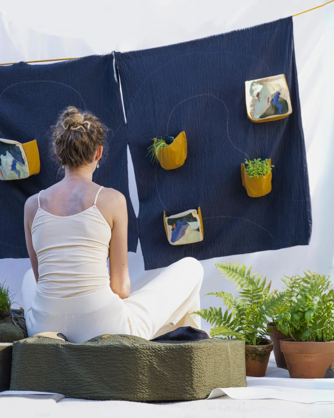 Alice Burnhope sitting on a cushion looking at her artwork 'A Sense of Nature' feauturing hand- stitched wall plants, hanging on a wall