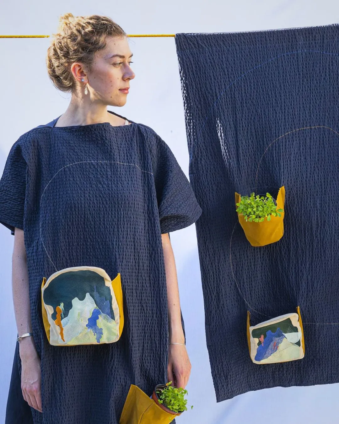 Alice Burnhope wearing a stitched artwork standing next to a textile 'wall' with stitched wall plants in pots.