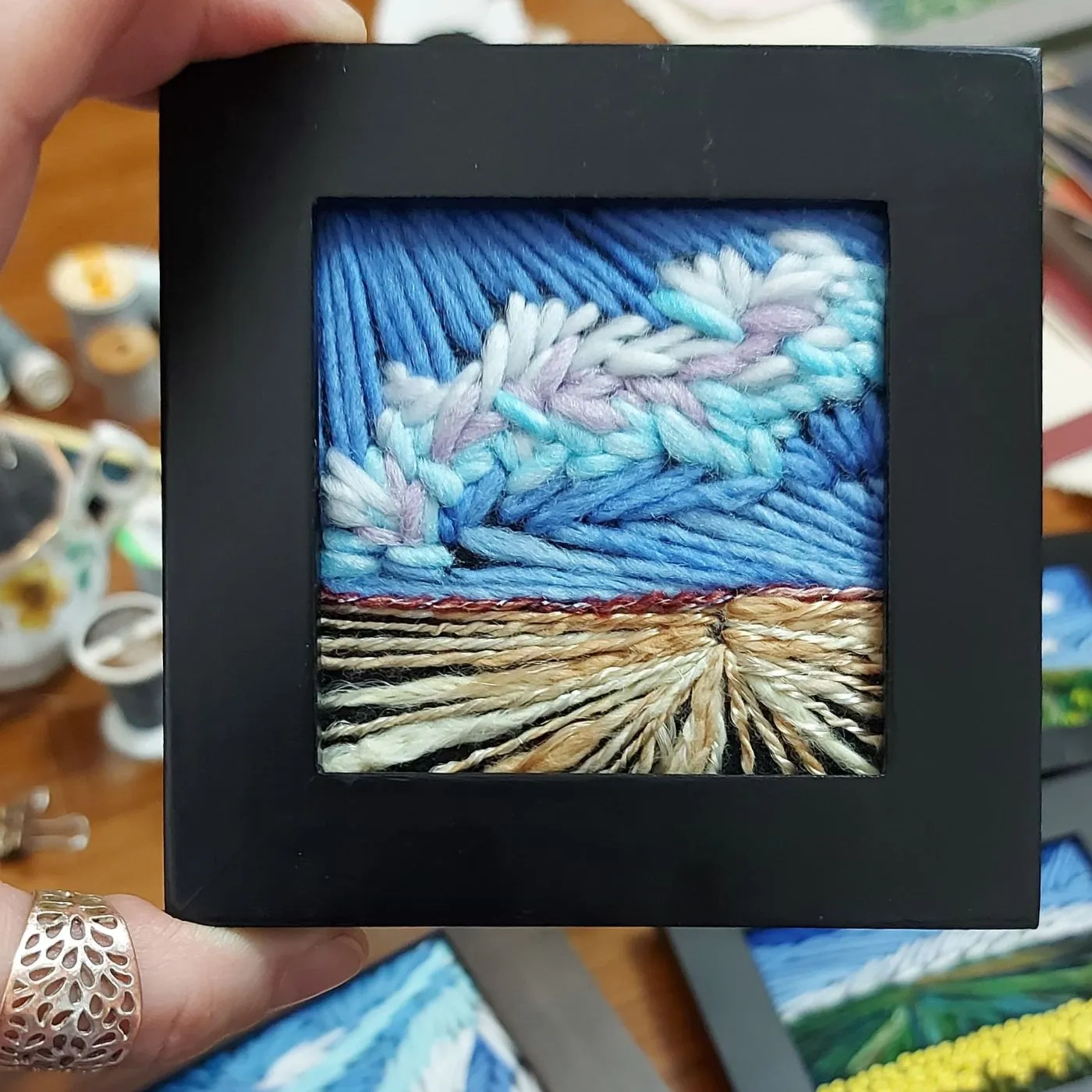 A hand holding a square picture of a stitched landscape, artwork. 