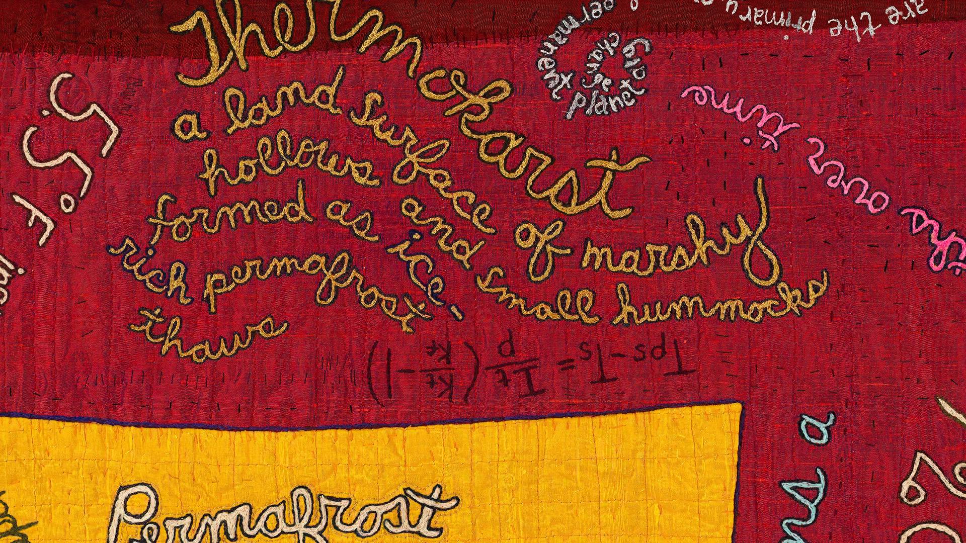 A close-up detail of a red and yellow patched art quilt with decorative handstitched writing across the fabric