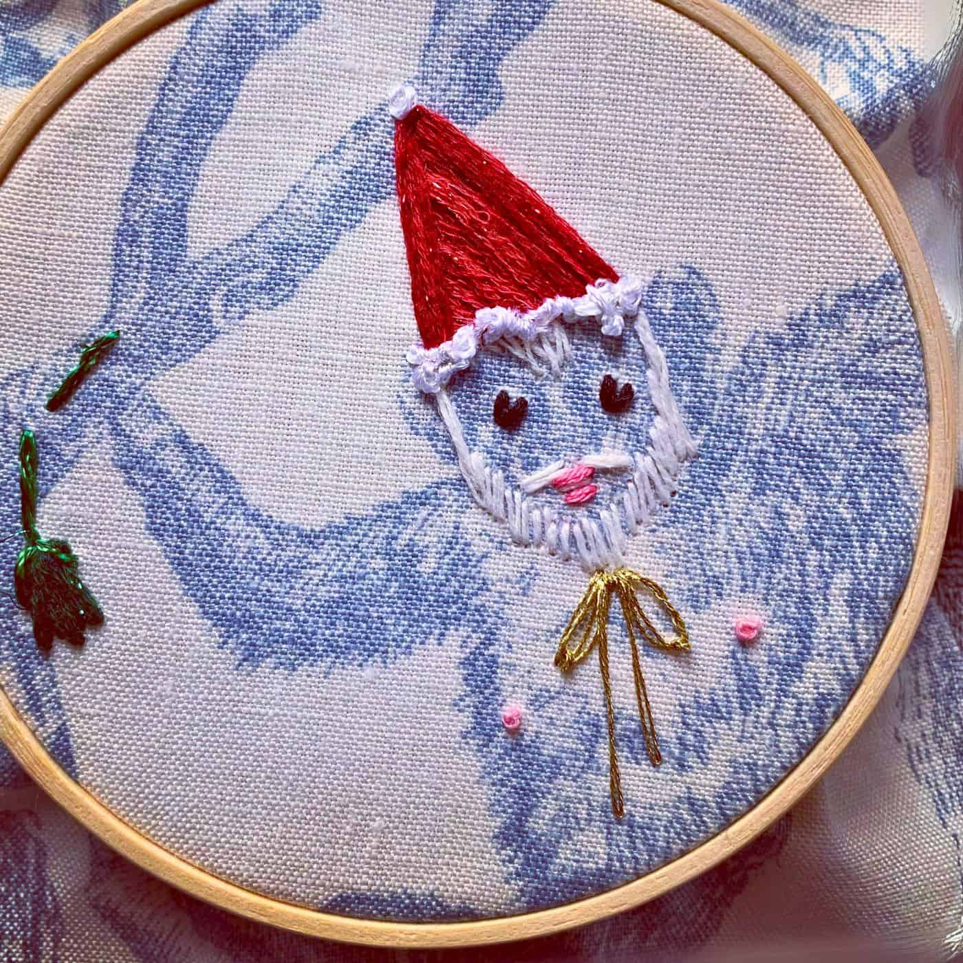 An embroidery hoop with a fabric image of a monkey. The monkey has a hand stitched santa hat and beard, a gold bow tie and his nipples and lips are stitched in pink thread.