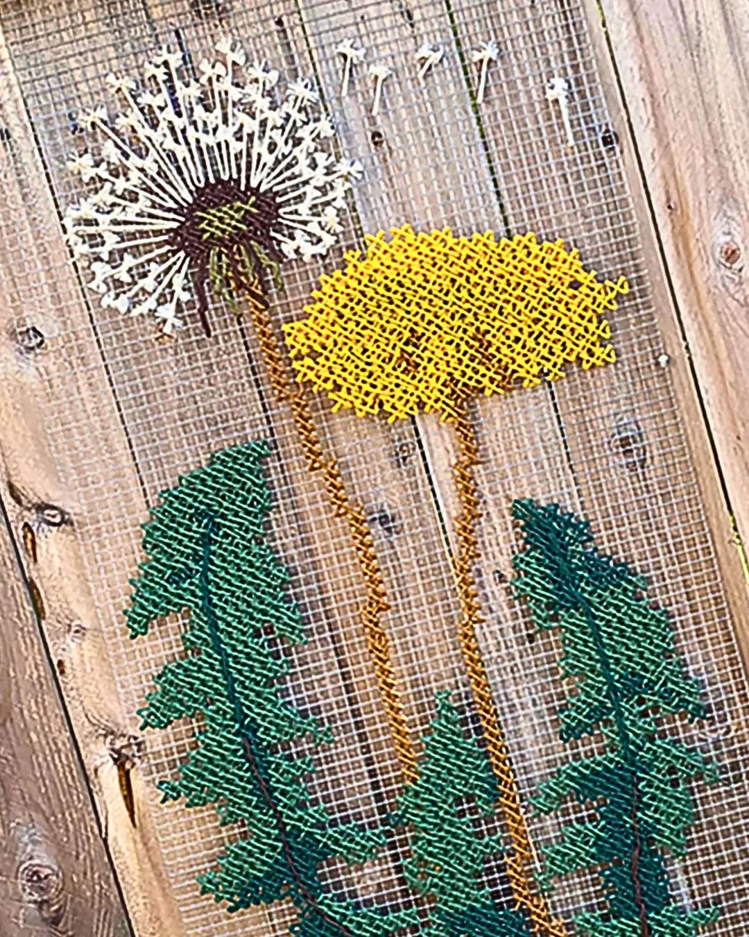 A hand embroidery artwork of a yellow dandelion with green leaves, stitched on to wire mesh and hung on a fence.