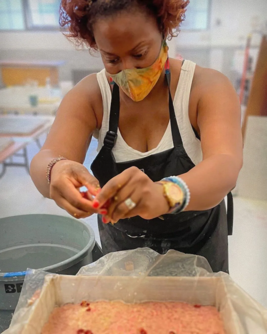 A woman wearing a protective mask and apron mixing a pink paste in her art studio