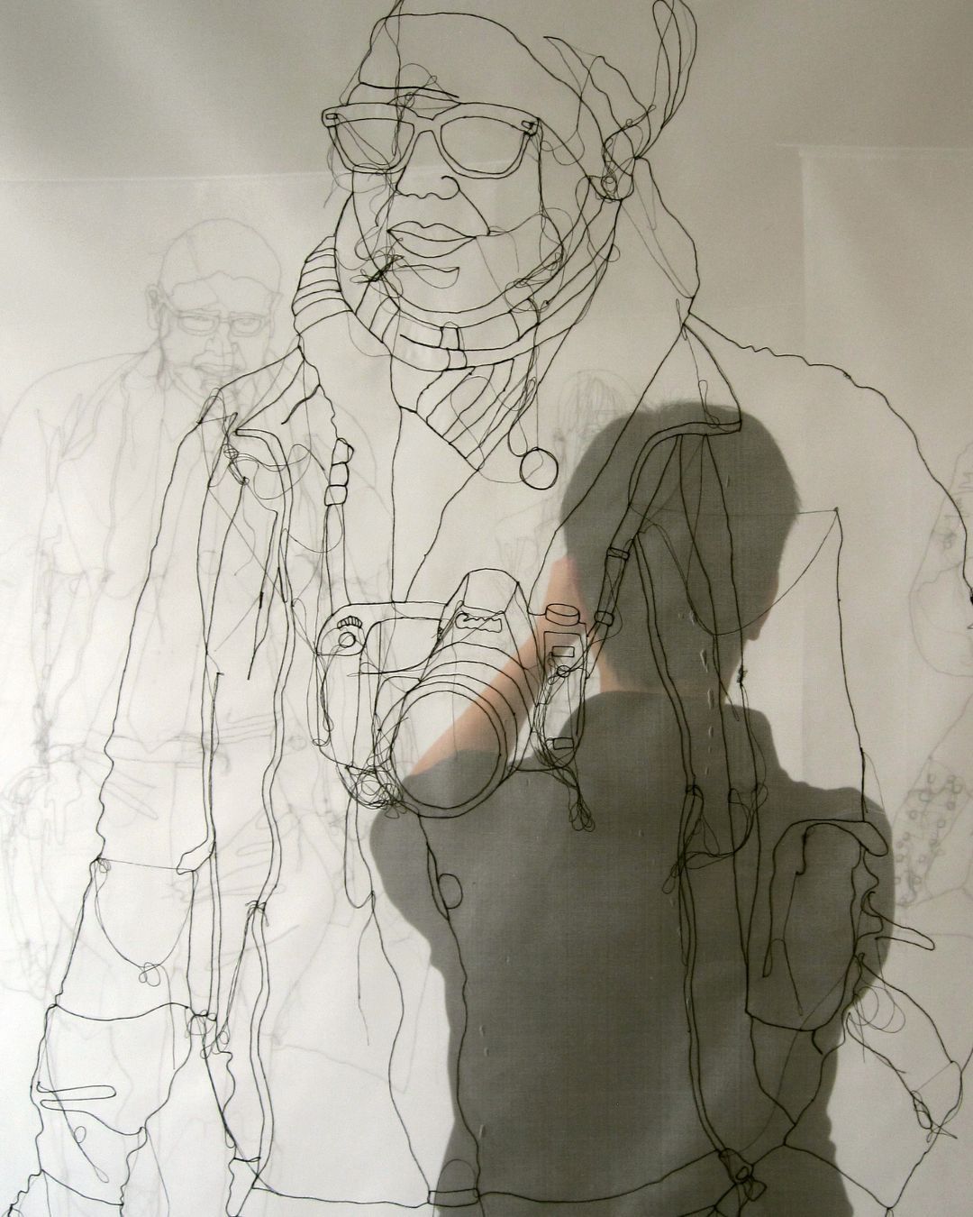 Stitched illustration of a winter-clad figure with sunglasses and camera, set on a semi-transparent background, subtly revealing the artist’s silhouette behind her work