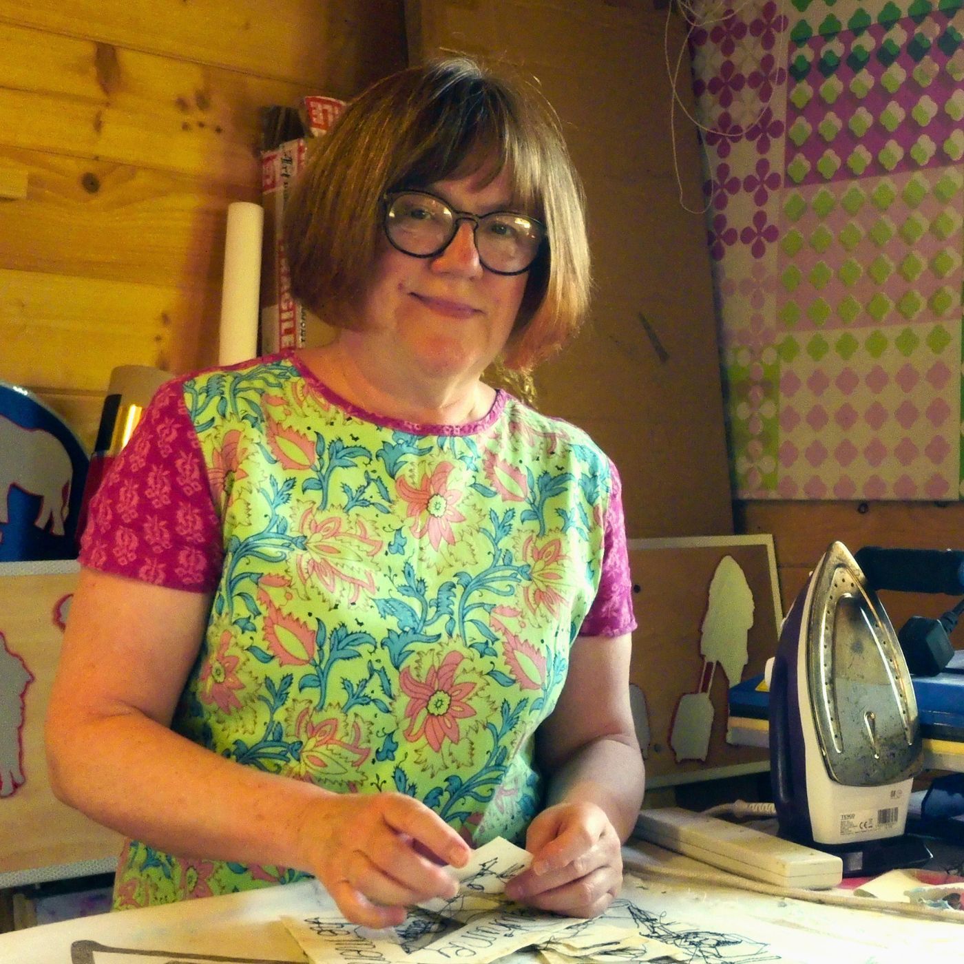 A female artist in glasses stood at her studio table handling small samples of her work 