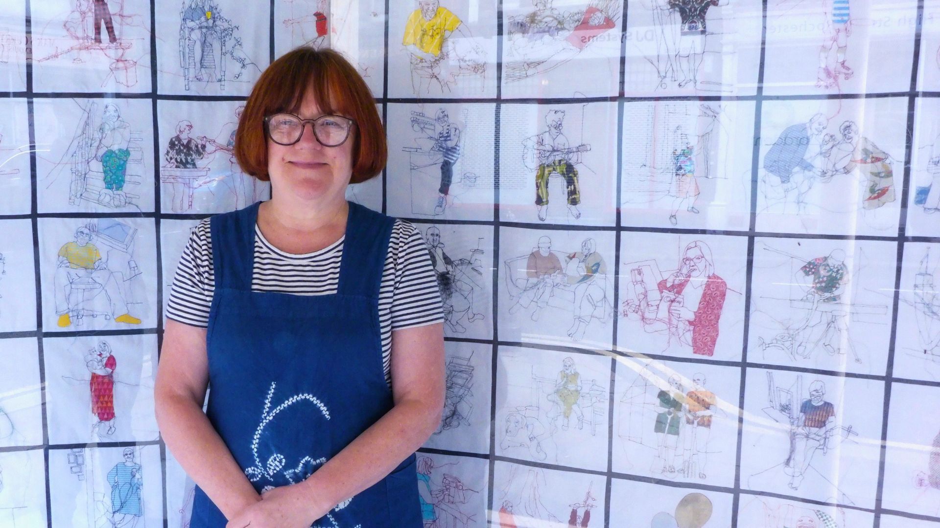 A female artist wearing glasses standing in front of a wall of her stitched drawings