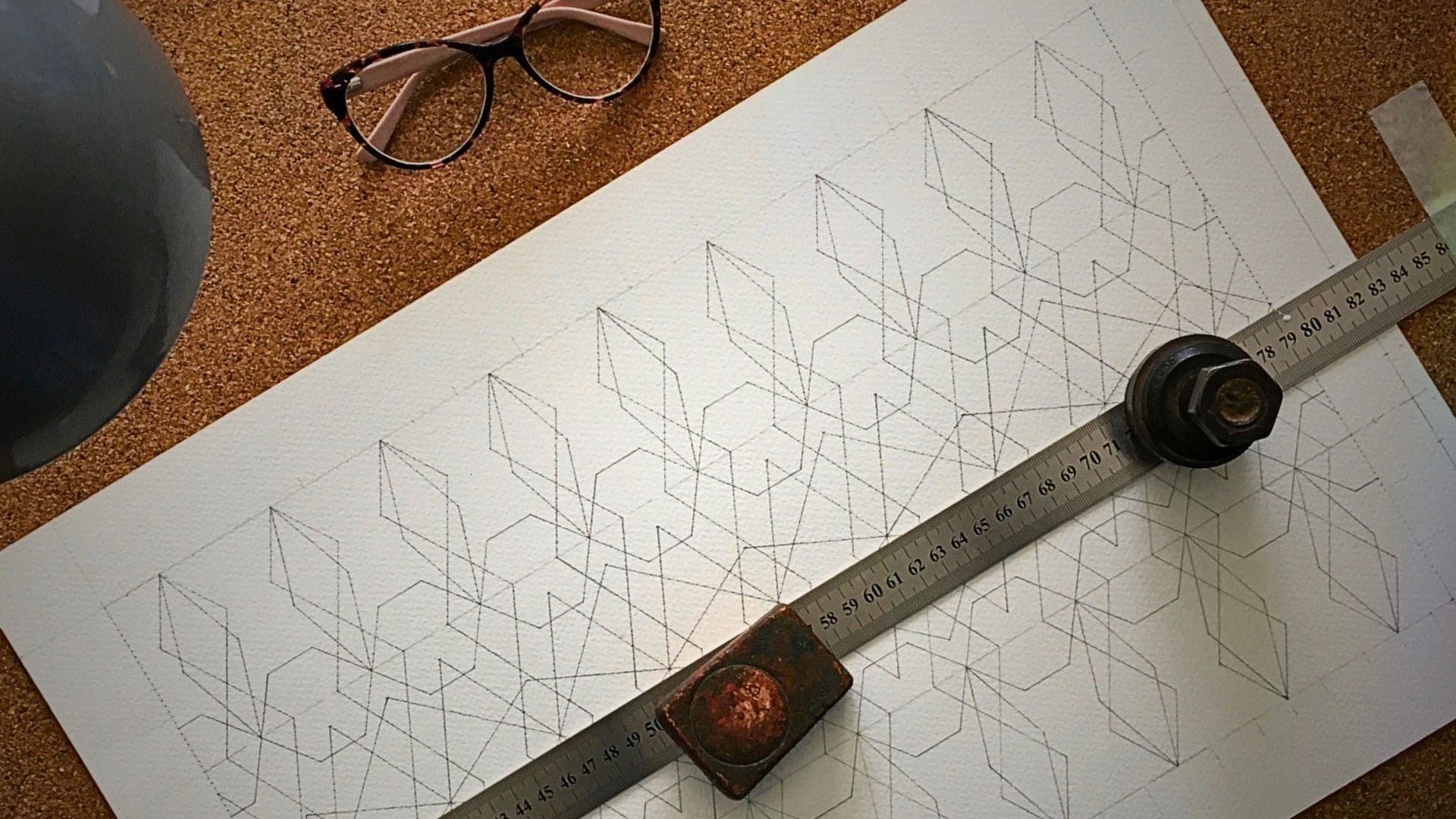 An image of a pattern being drawn on a piece of paper with a ruler and a pair of glasses on a cork board.