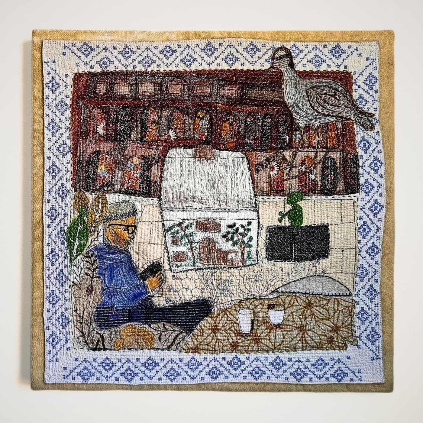 An abstract layered fabric artwork featuring a bird, a man sitting and a house in the middle.