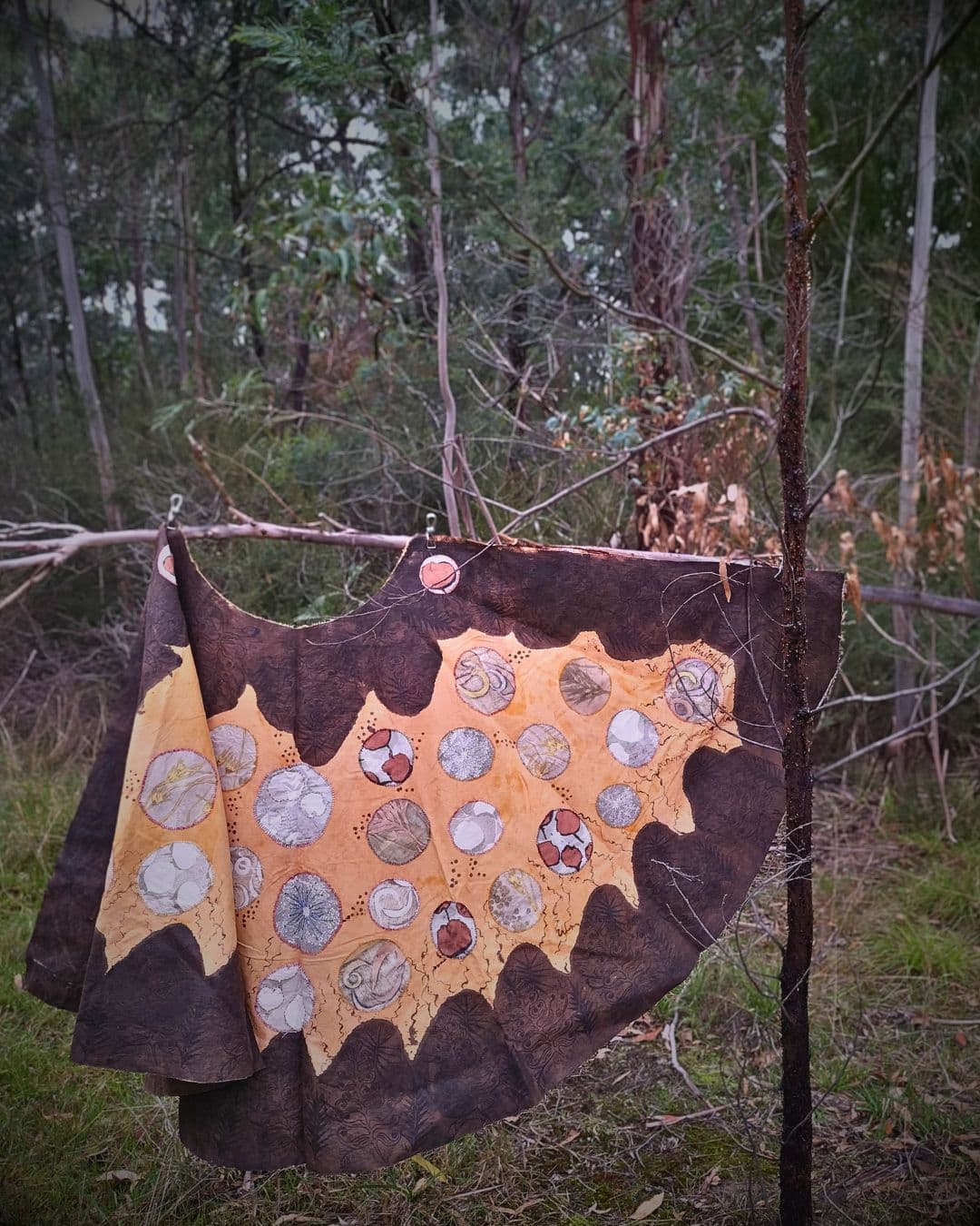 A stitched artwork displayed on a pole hanging in the woods.