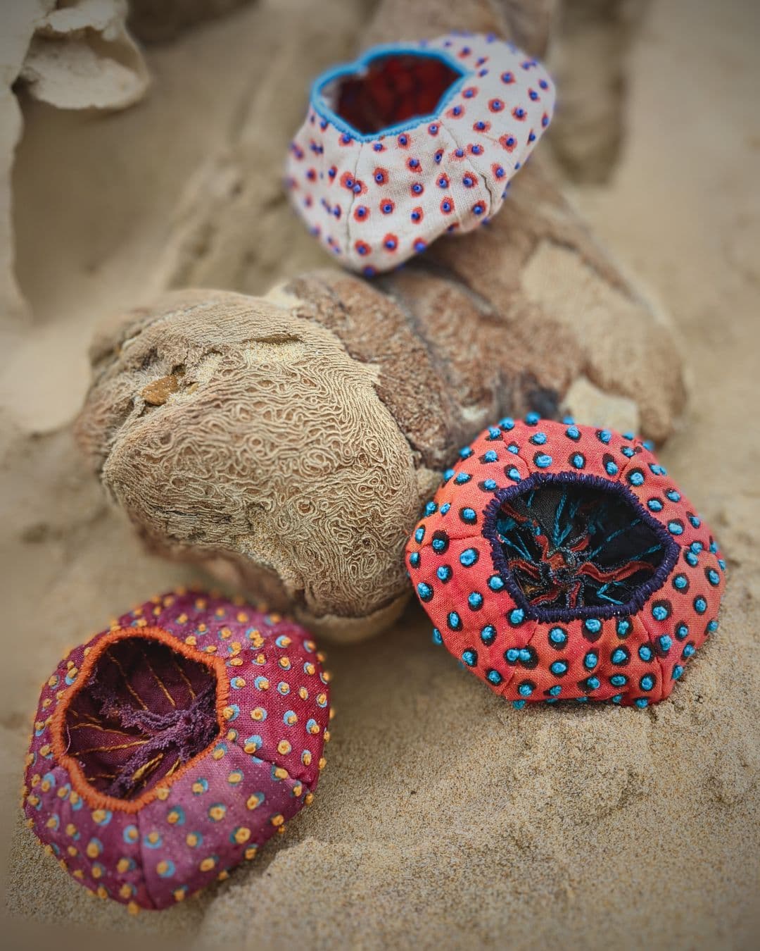 A group of colourful stitched 3D artworks objects on a sandy background.