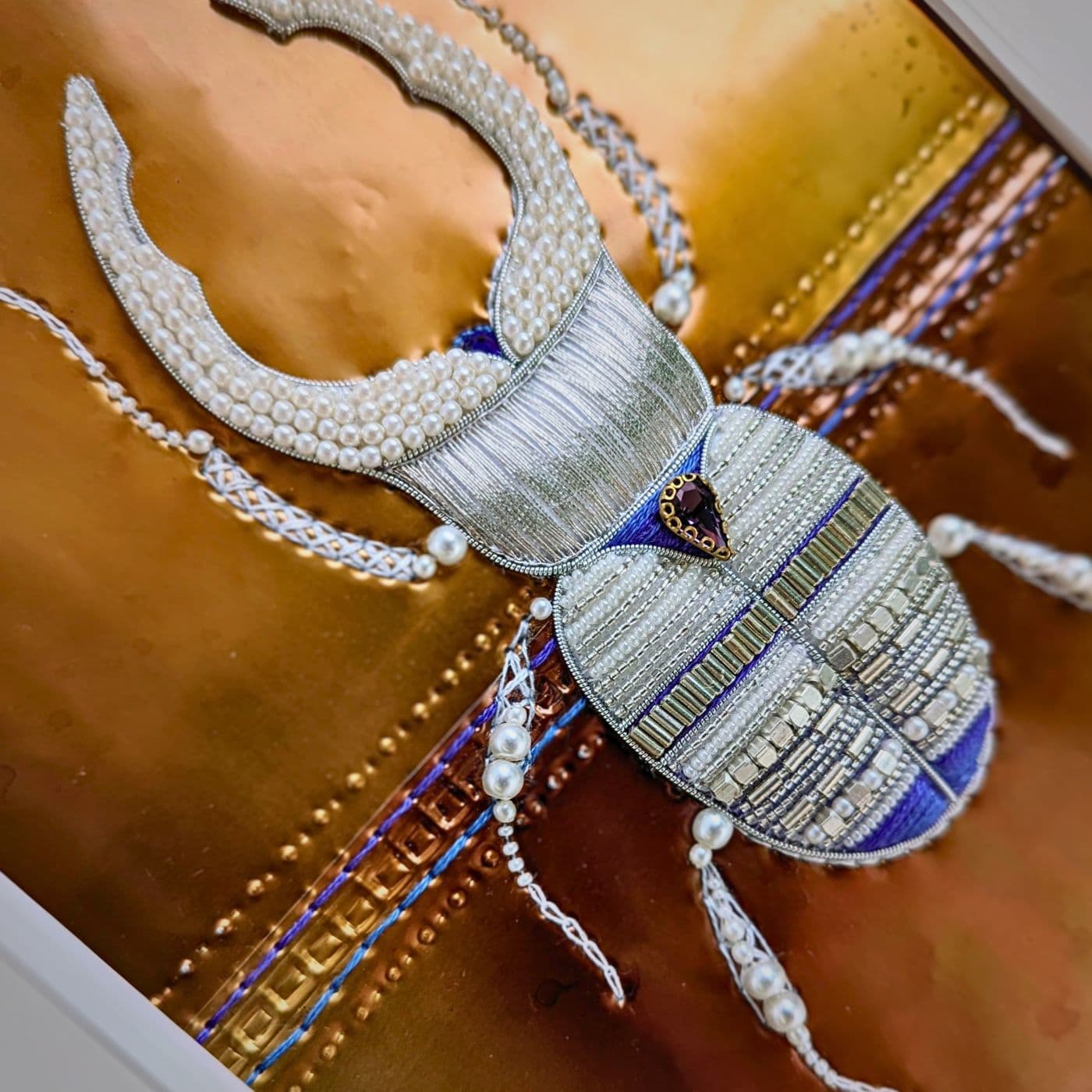 An art work of a male stag beetle embellished with beading and stitched on to a gold metal background. 