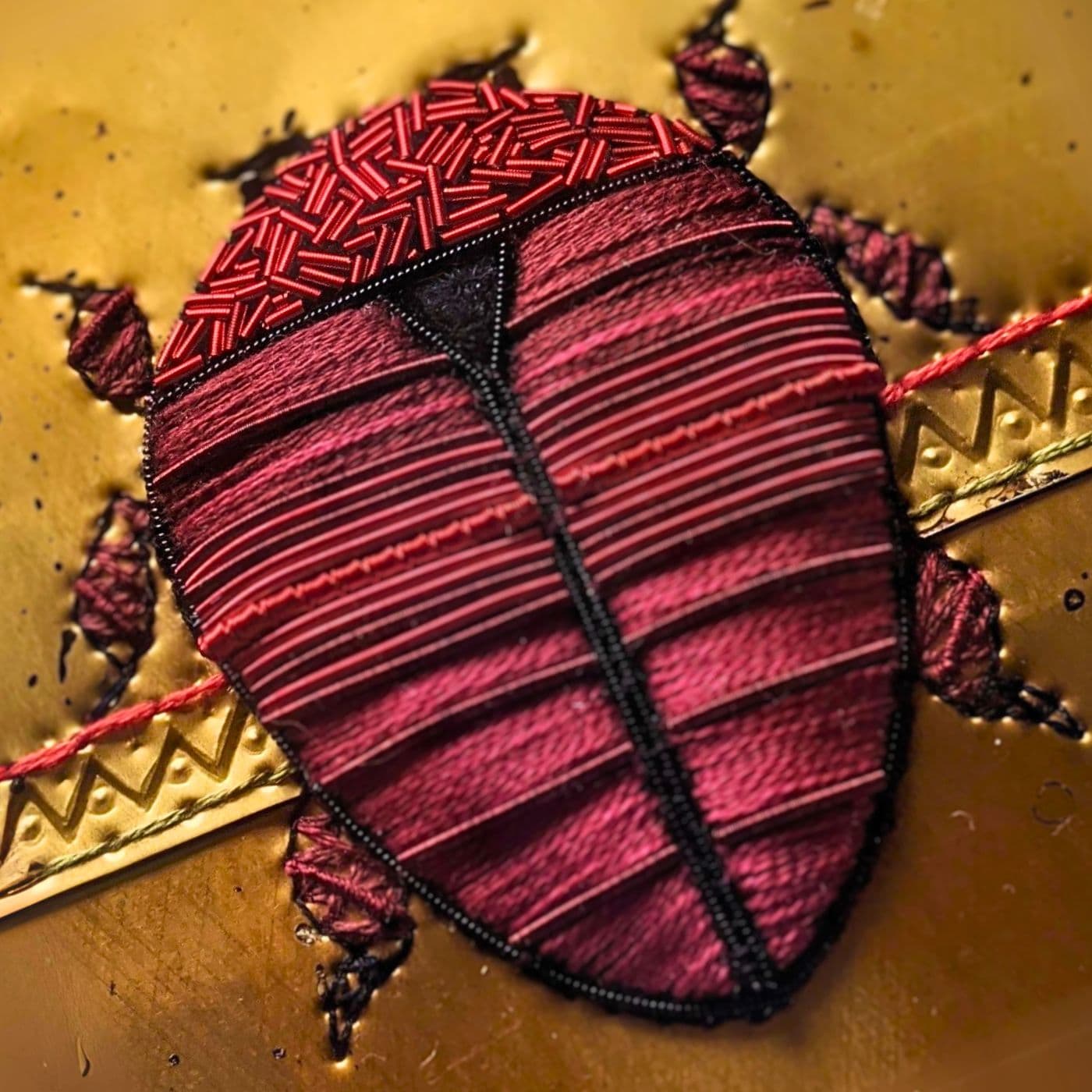 A close up of an art work of a red beetle stitched on to a gold metal surface. 