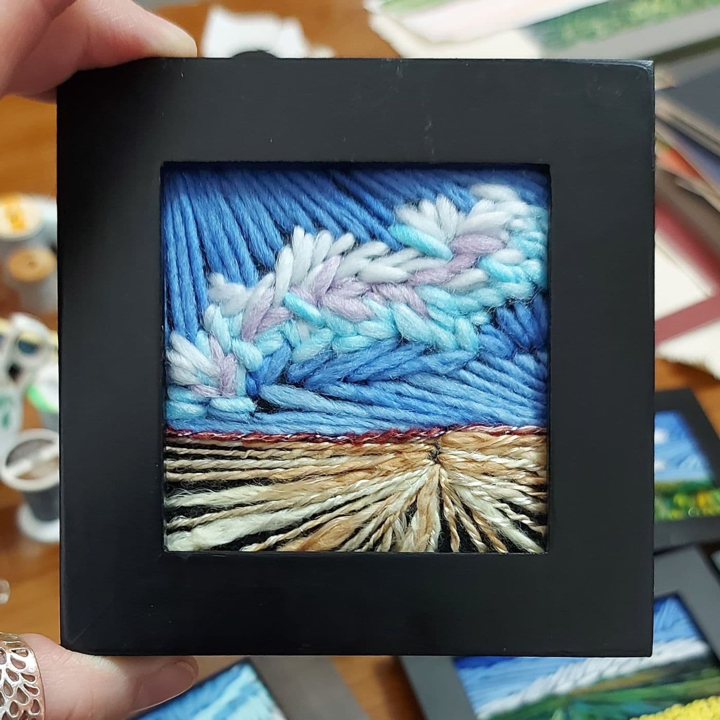 A hand holding a square picture of a stitched landscape, artwork. 