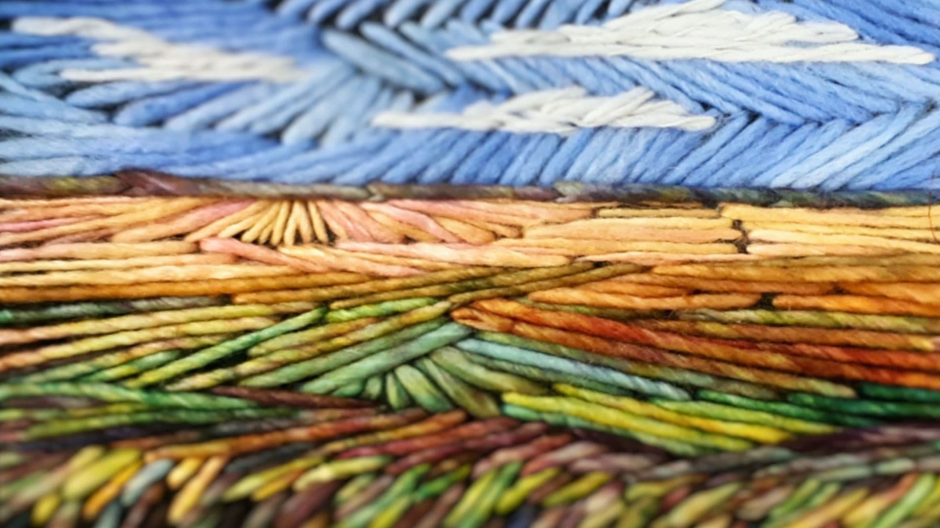 Close up of a colorful yarn artwork depicting an autumnal field with clouds in a blue sky