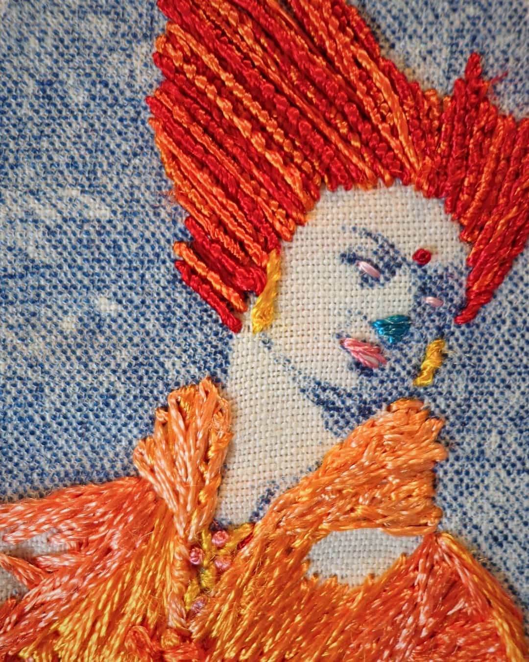 A close up of a person's face. Their hair is hand-stitched in bright orange thread and their clothing is a slightly lighter shade of orange. Their features are stitched in various bright colours.