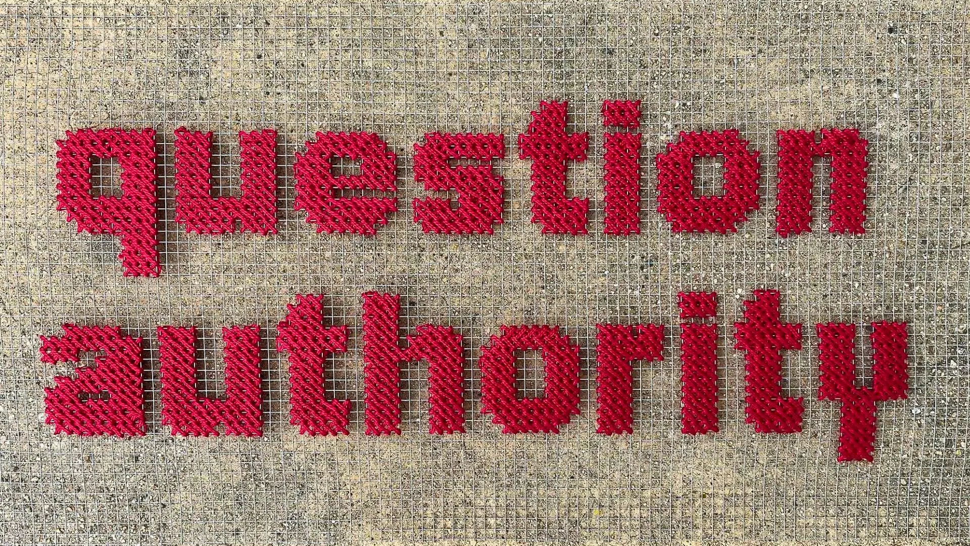 Red letters embroidered on to wired mesh, displayed on a stone background.