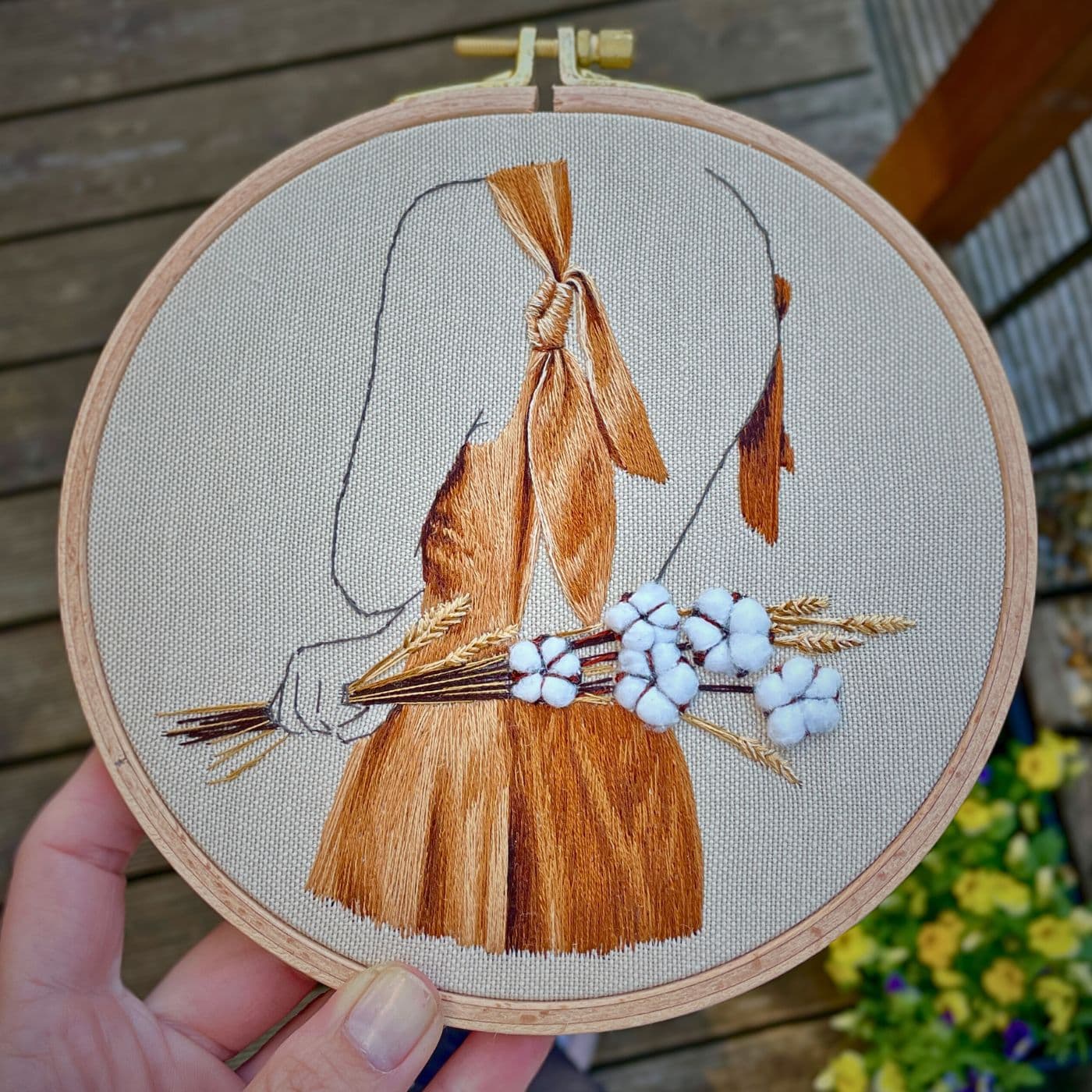 a hand holding an embroidery hoop with a stitched artwork of a girl holding a cotton bouquet. 