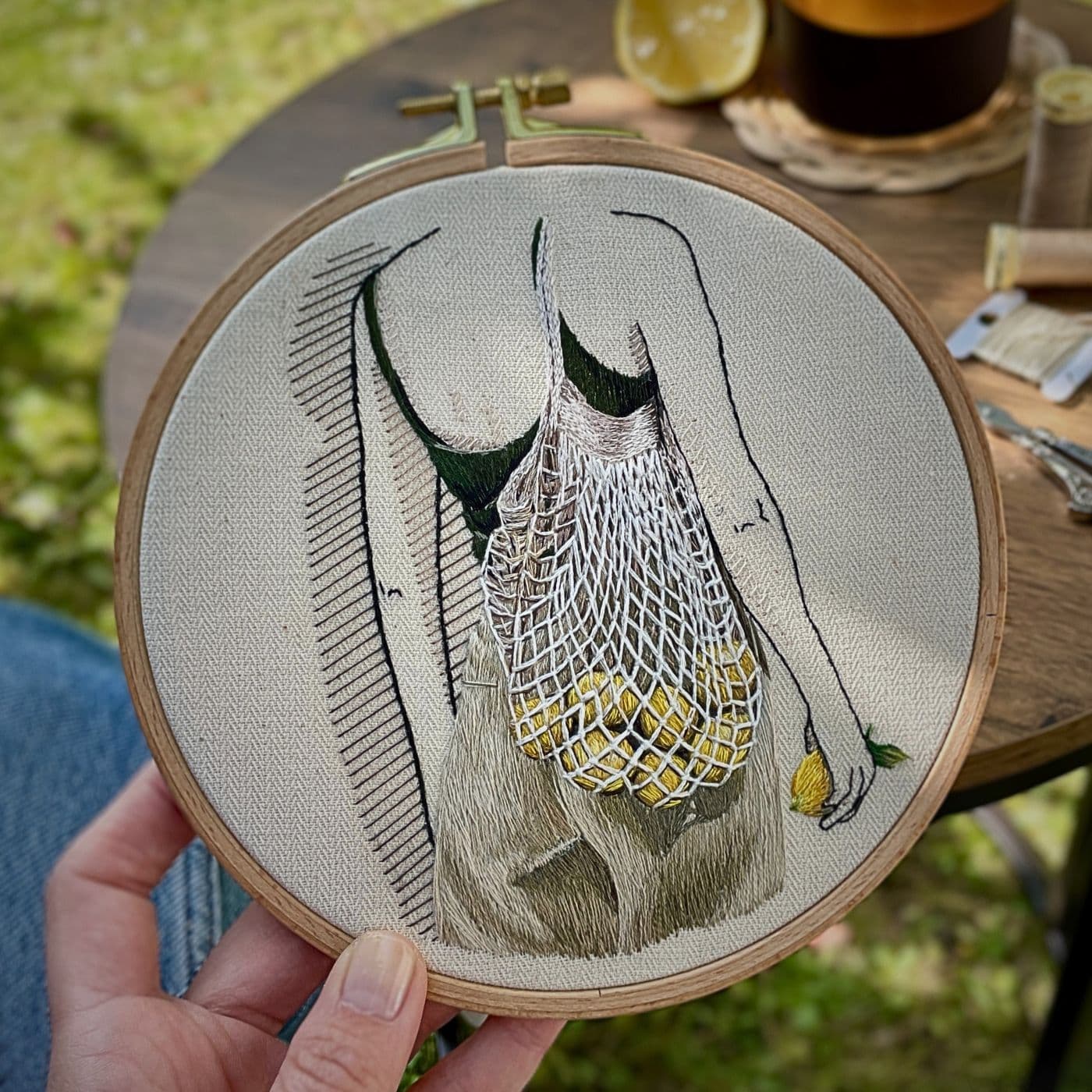 a hand holding a embroidery hoop with a stitched artwork of a woman holding a bag of lemons over her shoulder. 