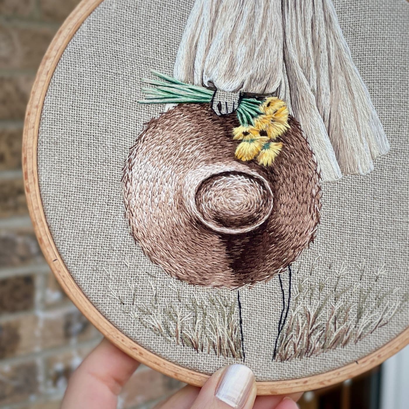 An embroidery hoop with a stitched artwork of a woman holding a hat and a bunch of daffodils. 