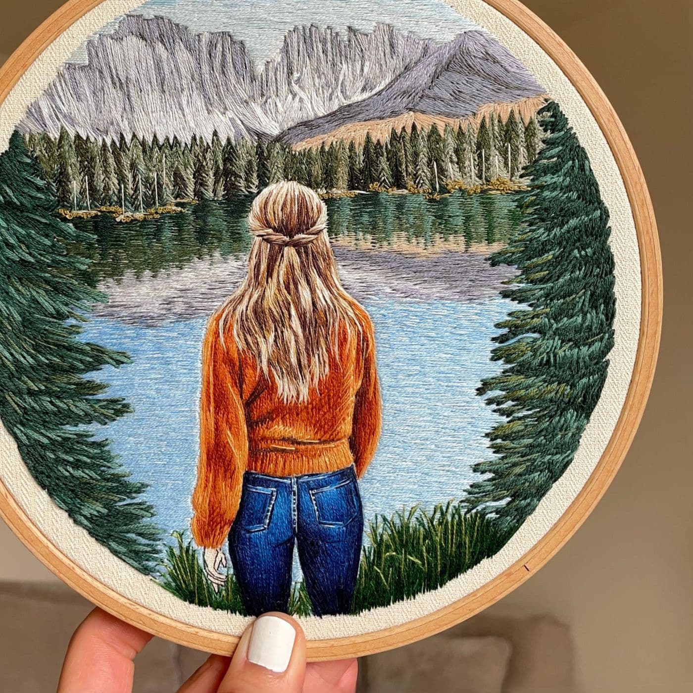 An embroidery hoop with a stitched artwork of a woman looking at a lake surrounded by trees and mountains.