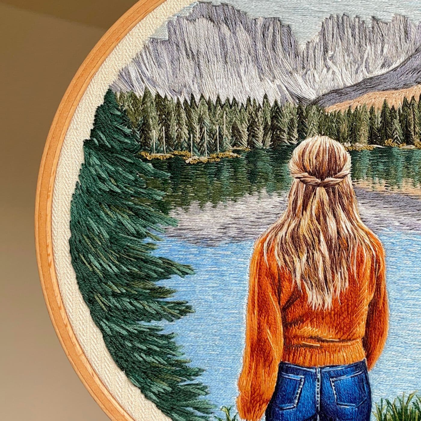 A close up of an embroidery hoop with a stitched artwork of a woman looking at a lake surrounded by trees and mountains.