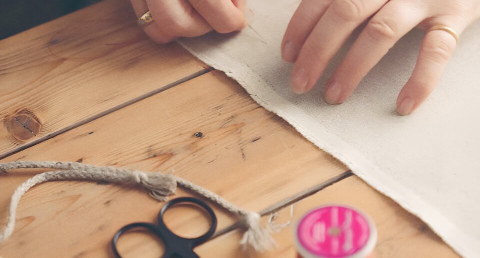 Close-up of hands delicately stitching fabric with a needle, surrounded by sewing tools on a wooden table, showcasing the artistry of textile creation